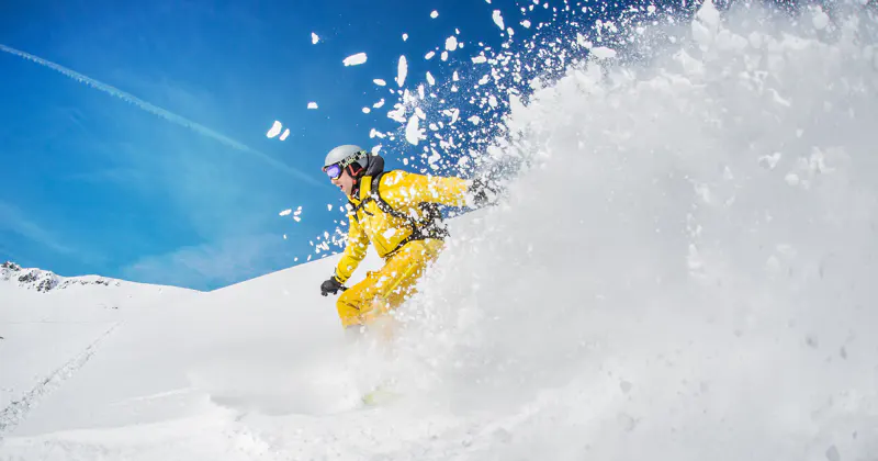 Een snowboarder in een geel jack glijdt door de verse sneeuw en laat een wolk poedersneeuw opstijgen tegen de helderblauwe lucht op een zonnige berghelling.